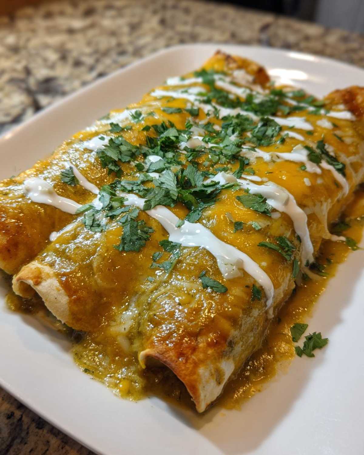Three rolled green chile chicken enchiladas covered in green sauce, melted cheddar cheese, sour cream drizzle, and cilantro.