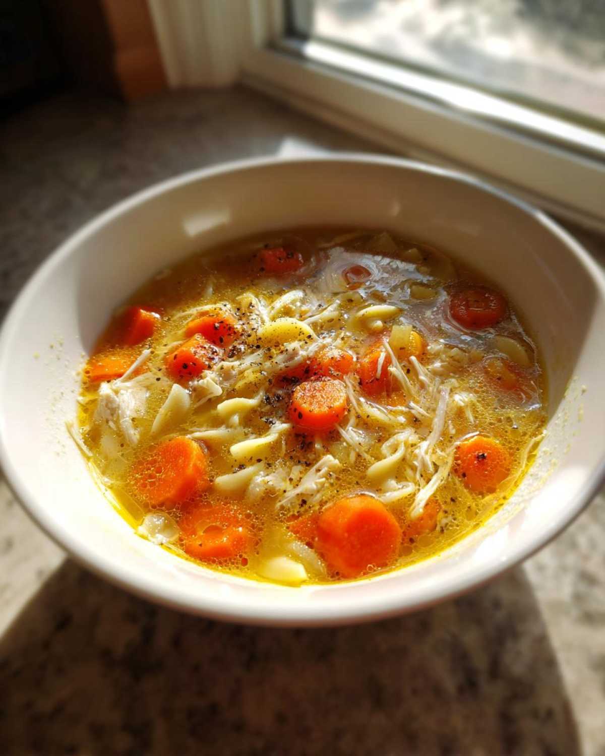 A close-up of a warm bowl of turkey soup with bright orange carrots, noodles, and black pepper seasoning.