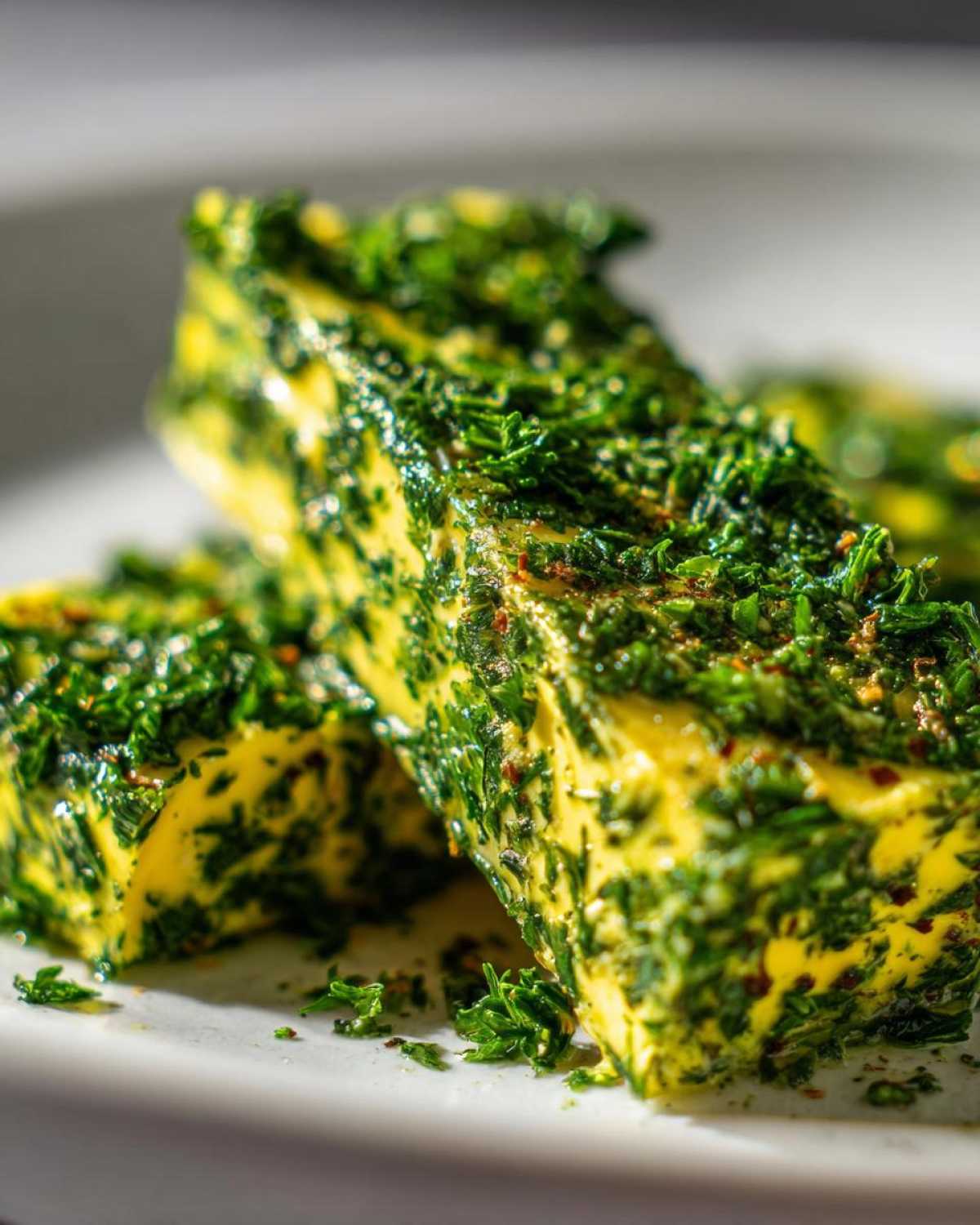 Close-up of two logs of bright yellow homemade garlic butter heavily coated in finely chopped green herbs.