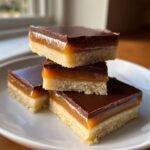 A stack of three homemade twix bar squares featuring shortbread, caramel, and chocolate topping on a white plate.