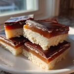 A stack of homemade twix bar squares featuring a shortbread base, caramel layer, and chocolate topping, sitting on a white plate.