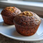 Close-up of two dark, richly baked honey bran muffins sitting on a white plate near a window.