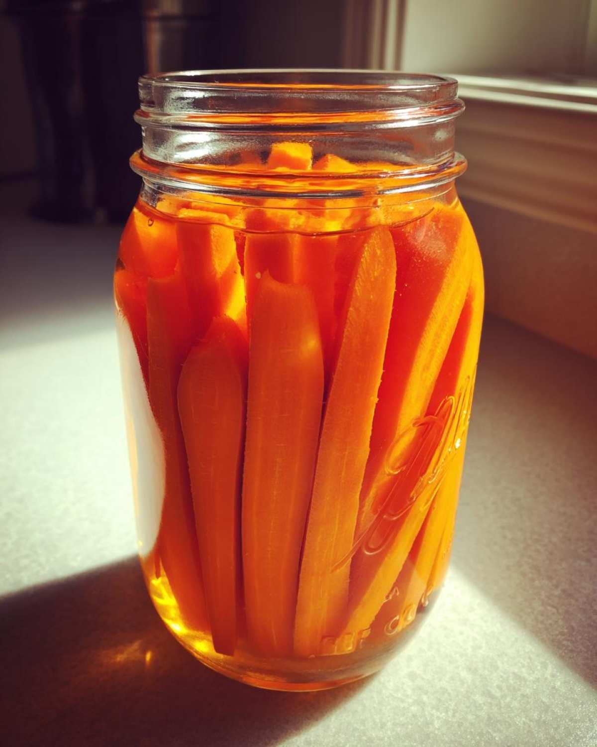 Close-up of bright orange carrot sticks tightly packed inside a glass jar submerged in pickling brine, ready for pickled carrots.
