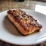 A close-up of a juicy, glazed air fryer salmon fillet resting on a white plate.