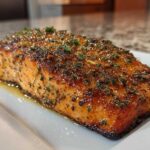 Close-up of a juicy, seasoned air fryer salmon fillet resting on a white rectangular plate with melted butter.