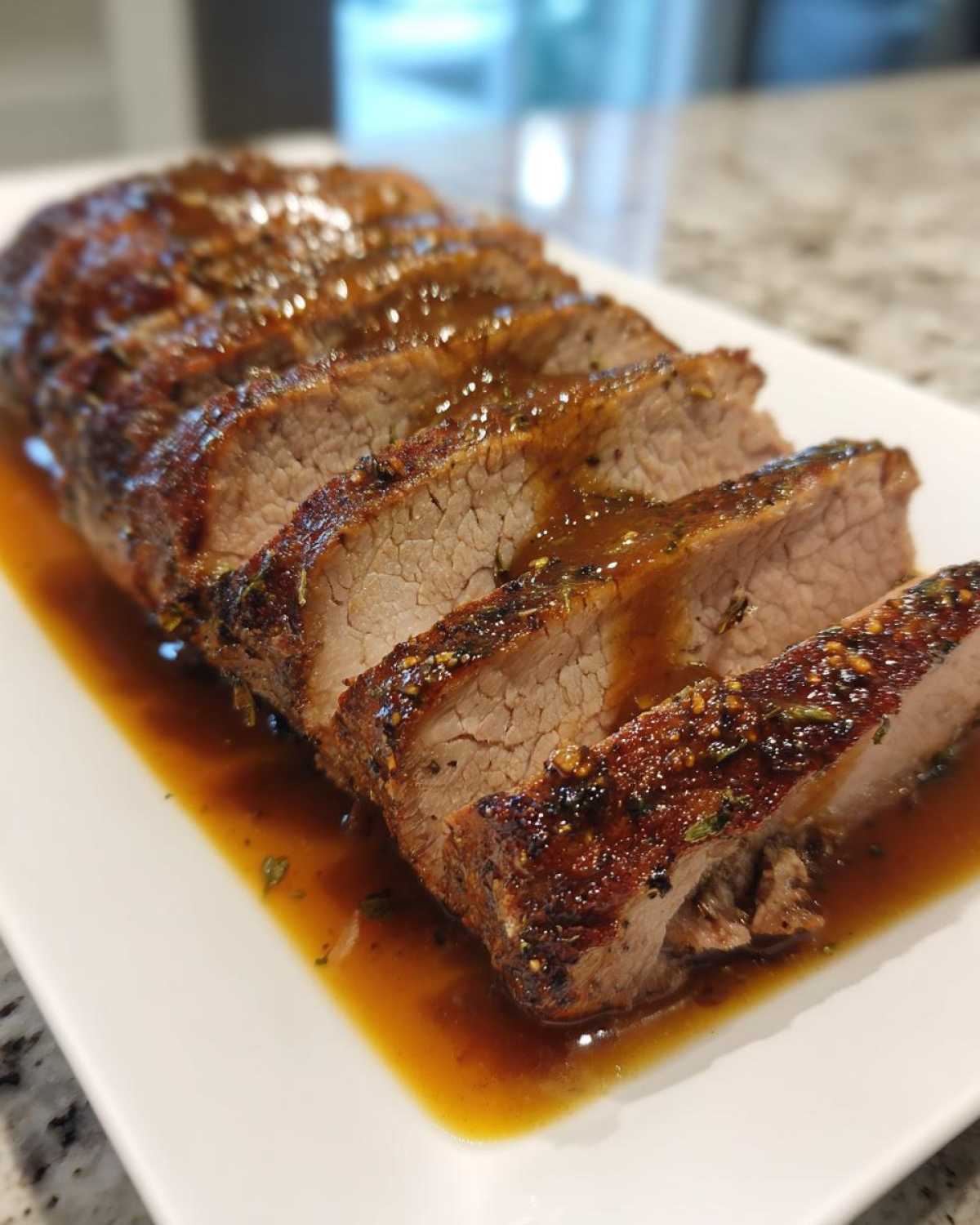 Close-up of a sliced pork loin roast, glistening with a rich brown glaze, resting in its own juices on a white platter.
