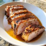 Close-up of perfectly cooked, sliced pork loin roast resting in its juices on a white plate.