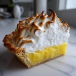 A close-up of a slice of lemon meringue pie showing the bright yellow filling, flaky crust, and tall, torched meringue topping.