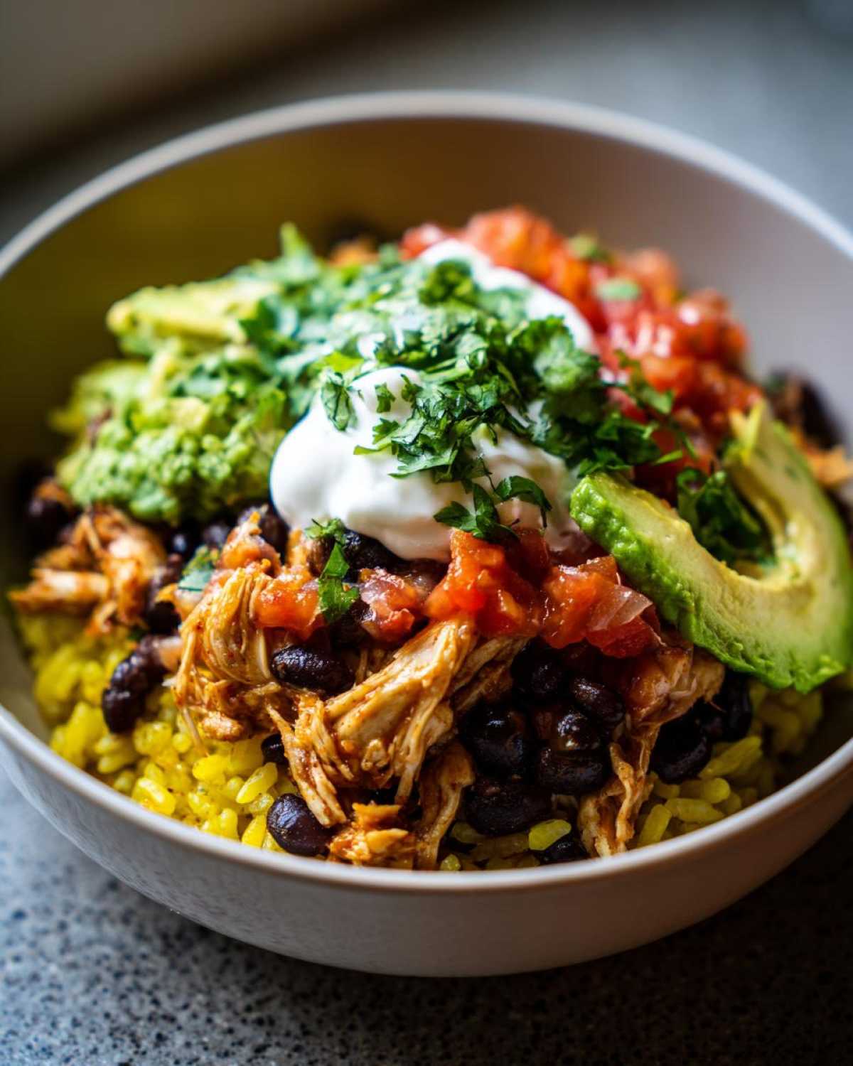 Close-up of a delicious chicken burrito bowl featuring shredded chicken, black beans, yellow rice, guacamole, sour cream, and salsa.