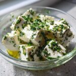 Cubes of white cheese soaking in oil and heavily topped with chopped green chives, ready for serving as marinated cheese.
