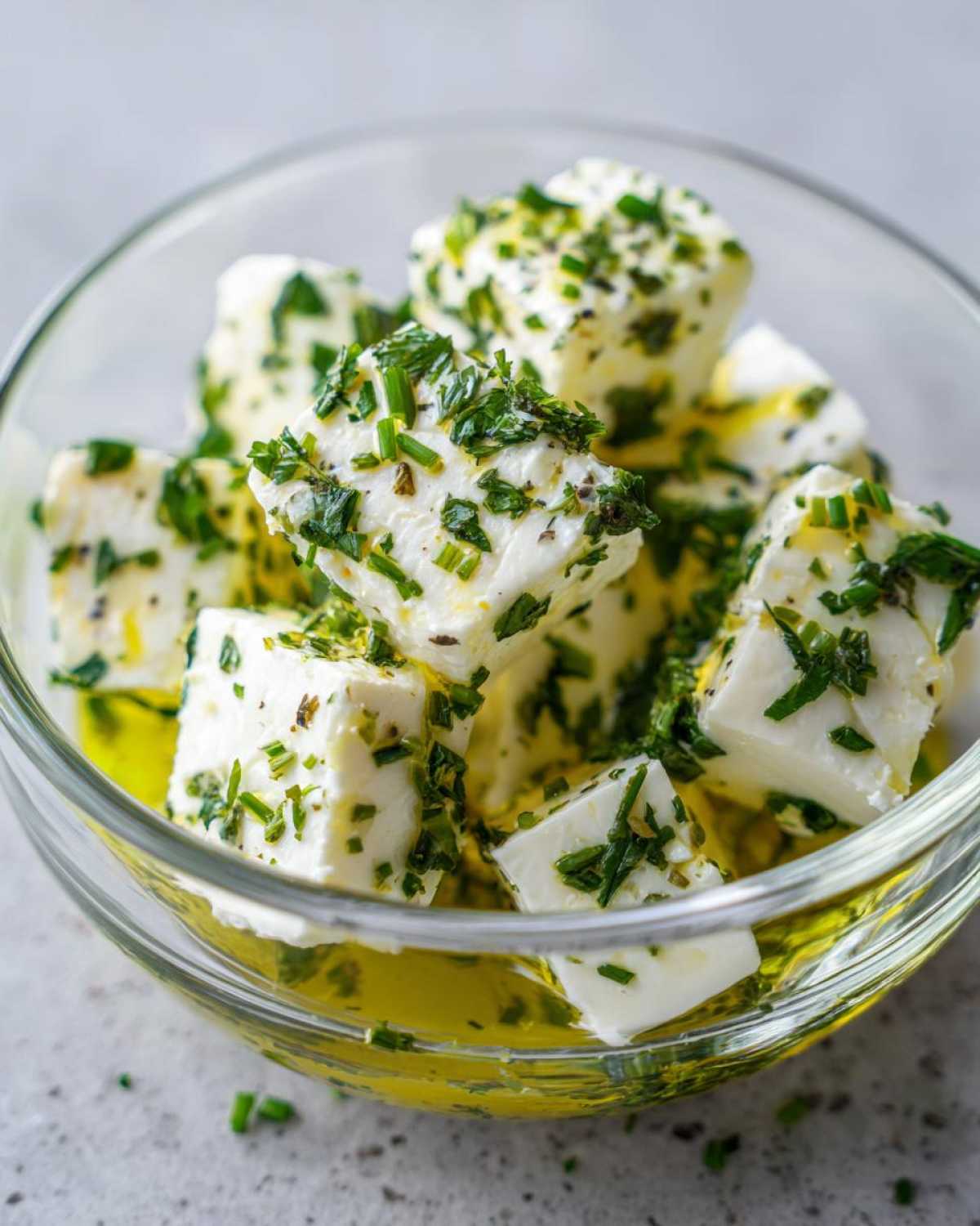 Close-up of white cheese cubes soaking in olive oil and heavily coated with fresh green herbs, ready for serving as marinated cheese.