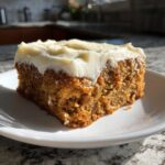 A close-up of a moist slice of applesauce cake topped with thick white frosting on a white plate.