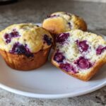 Three moist blueberry protein muffins, one cut in half showing the fluffy interior and berries.