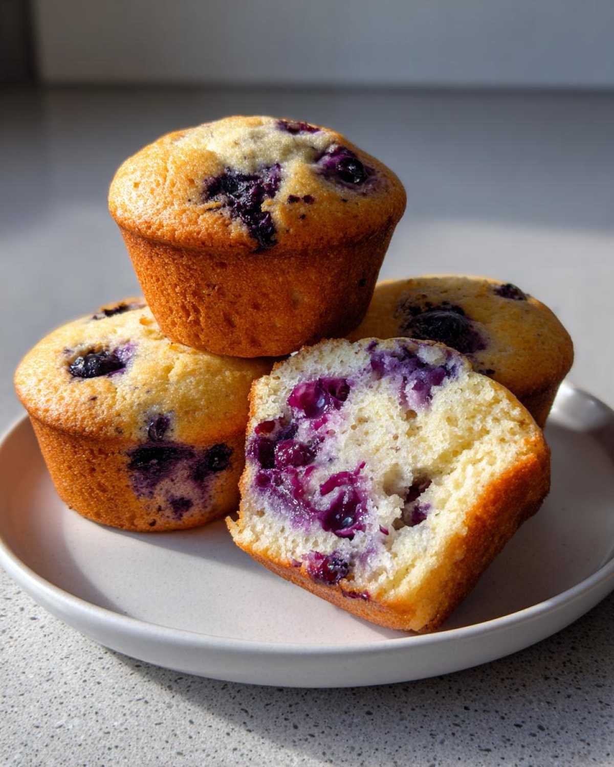 A stack of moist blueberry protein muffins, with one cut in half showing the soft interior and juicy berries.