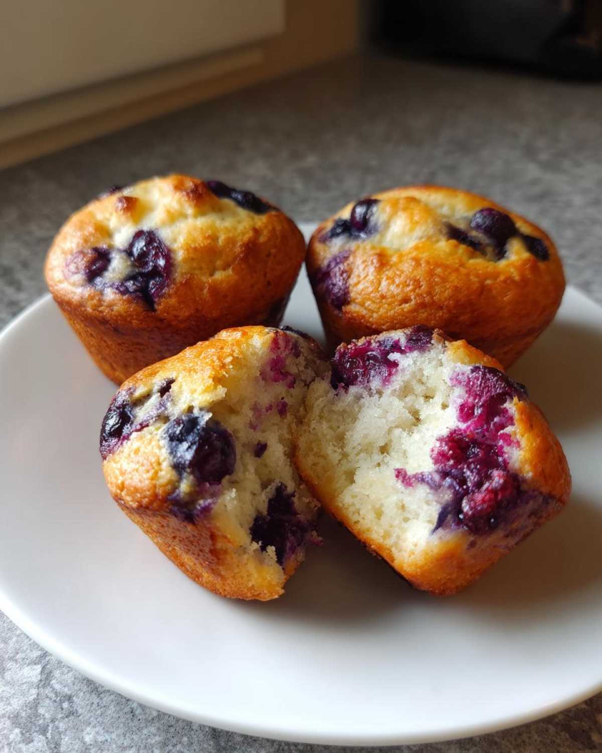 Close-up of four golden blueberry protein muffins, one broken in half showing the moist interior.