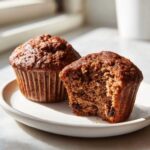 Two moist bran muffins on a plate, one bitten into, showing the texture and raisins inside.