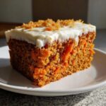Close-up of a moist slice of carrot cake recipe topped with thick cream cheese frosting and shredded carrot garnish.