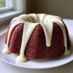 A moist red velvet bundt cake topped with thick, dripping cream cheese glaze on a white plate.