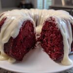 A moist red velvet bundt cake, cut in half, generously topped with dripping white cream cheese glaze.
