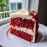 A perfect slice of moist red velvet cake layered with cream cheese frosting, sitting on a white plate.