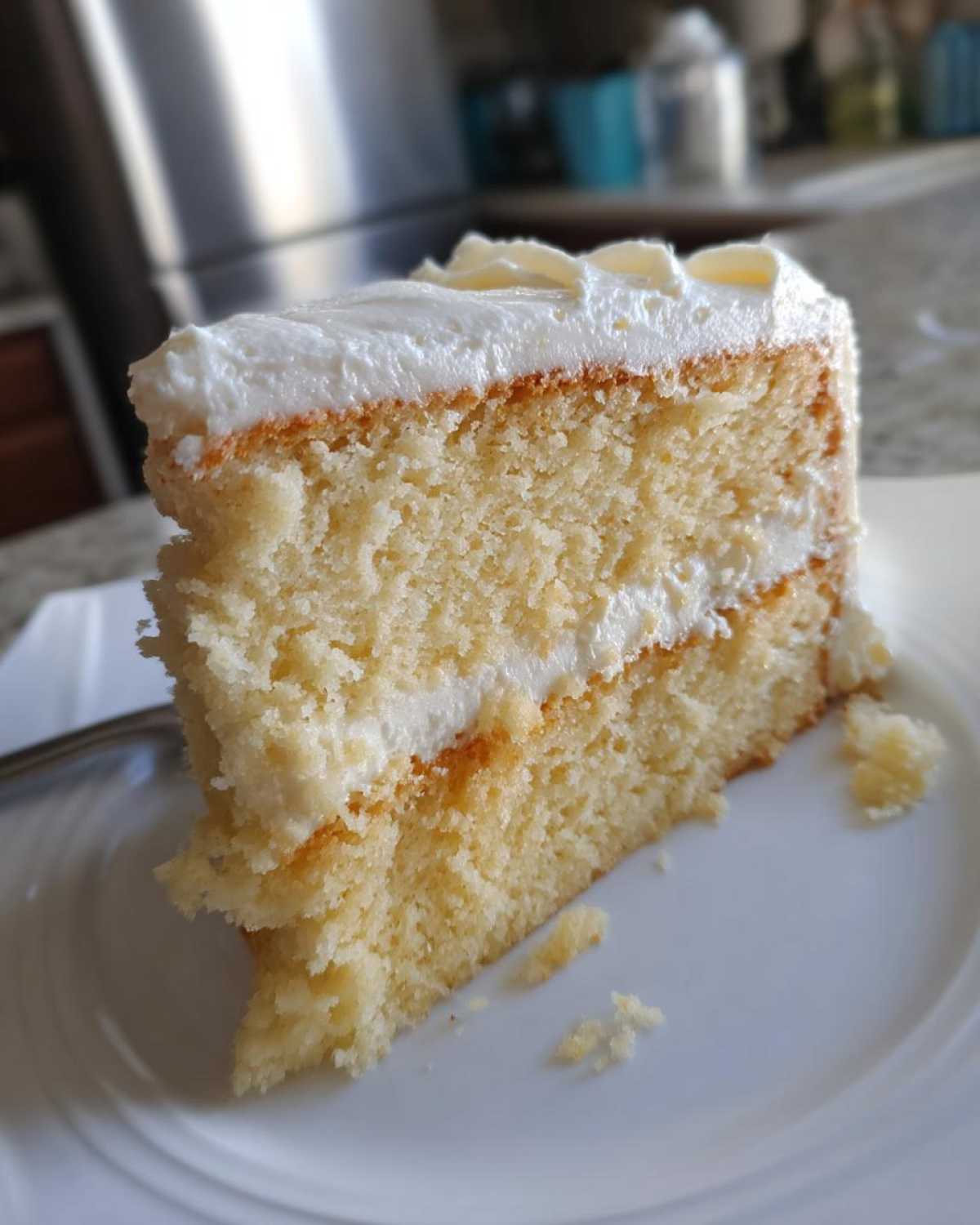 Close-up of a moist, two-layer slice of vanilla cake recipe with white frosting on a white plate.