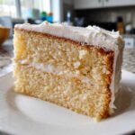 Close-up of a moist slice of vanilla cake recipe with thick white frosting layers.