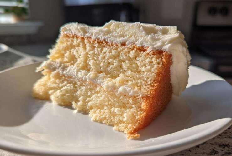 Close-up of a moist slice of vanilla cake recipe with thick white frosting, catching bright sunlight.