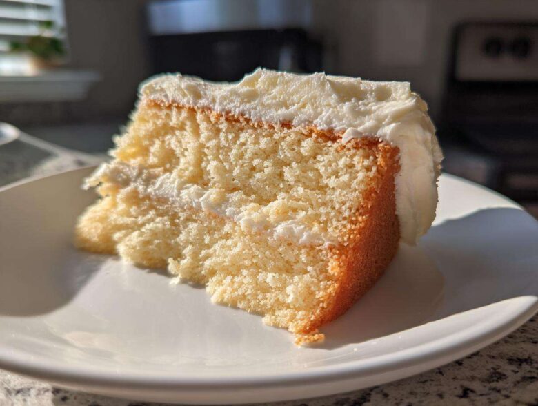 Close-up of a moist slice of vanilla cake recipe with thick white frosting, catching bright sunlight.