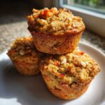 Three freshly baked morning glory muffins stacked on a white plate, topped with nuts and shredded carrots.