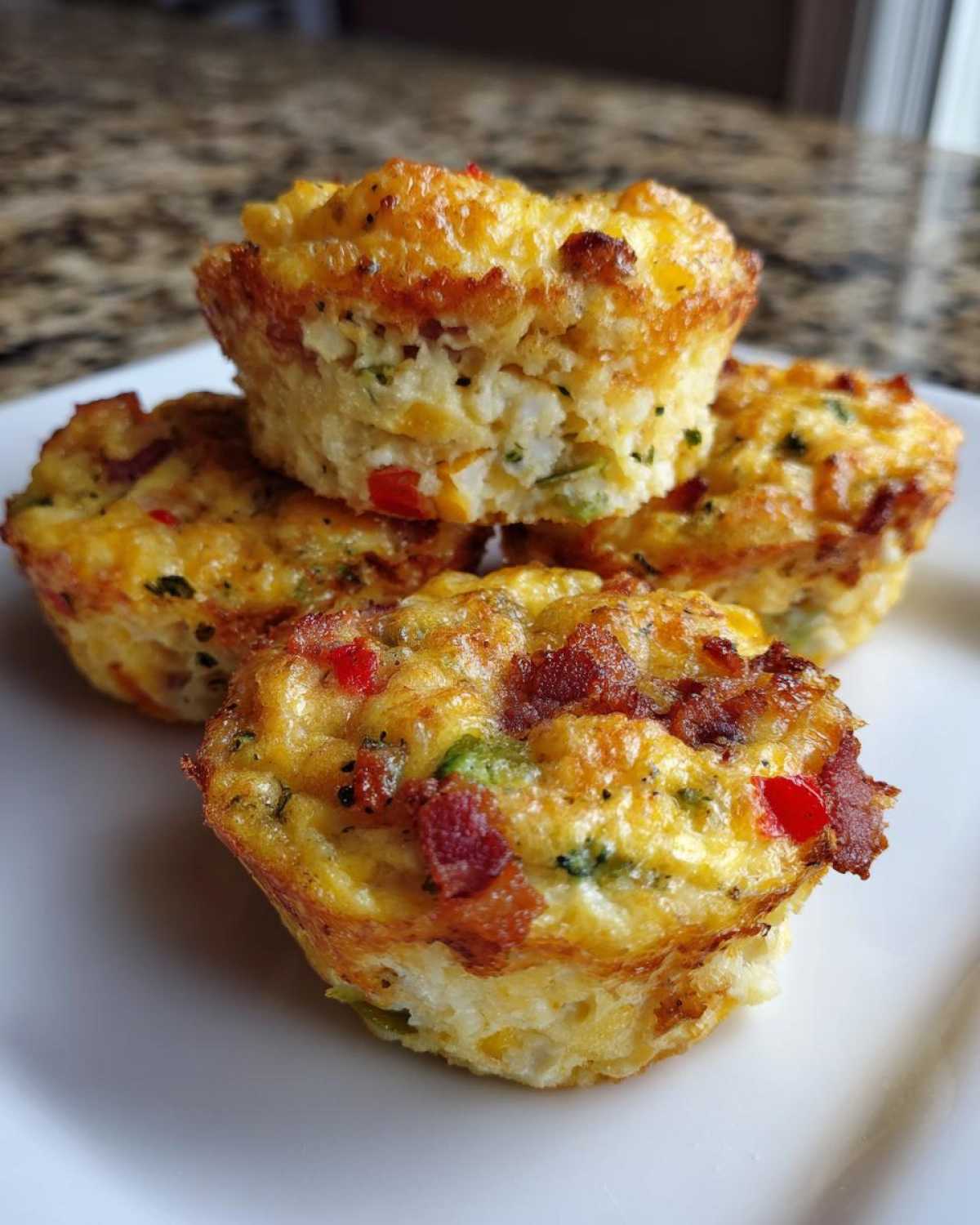 A stack of four golden brown muffin tin egg bites loaded with bacon and vegetables on a white plate.