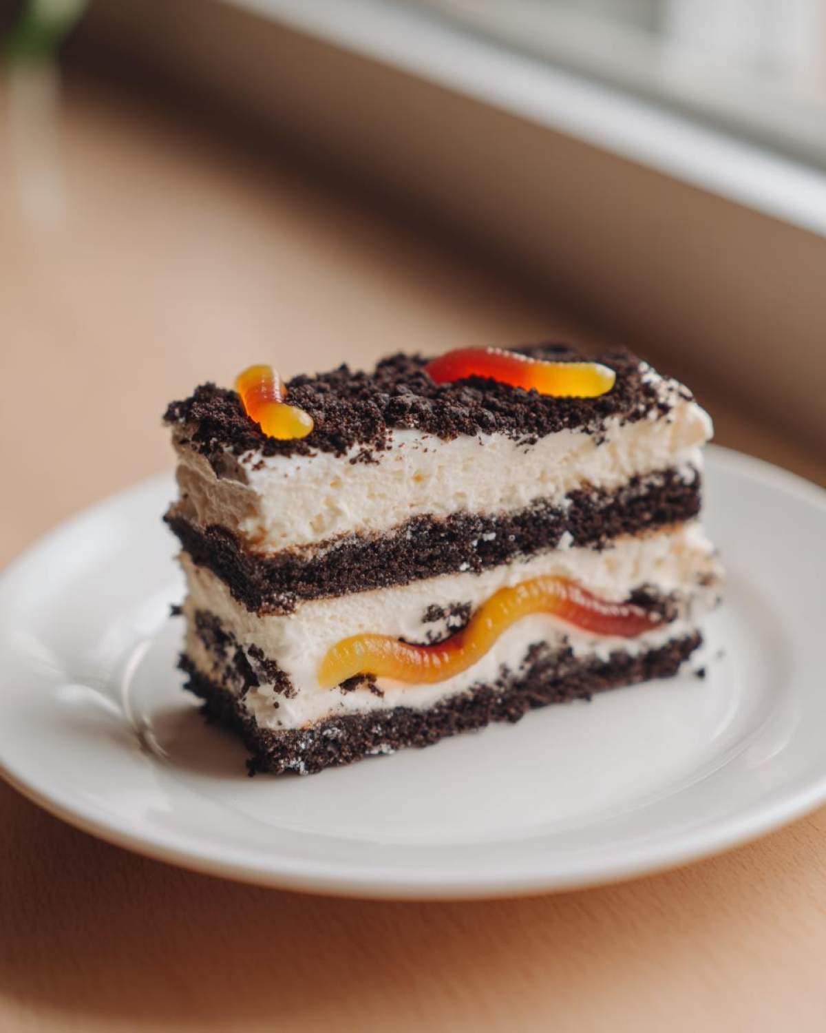 A layered slice of no-bake dirt cake featuring dark cookie crumbs, white cream filling, and gummy worms.