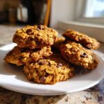 A stack of golden brown, oatmeal chocolate chip breakfast cookie piled on a white plate near a window.