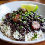 A close-up of a white bowl filled with white rice topped generously with seasoned black beans and rice and sliced sausage.