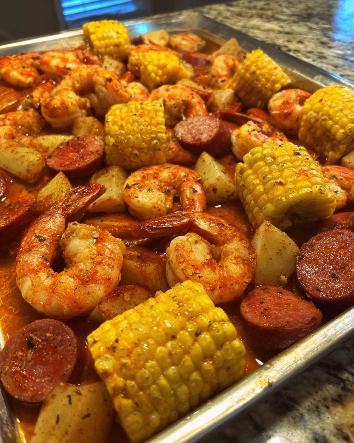 Close-up of a metal tray filled with seasoned shrimp, corn on the cob, potatoes, and sausage from an oven shrimp boil.
