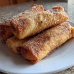 Four golden brown peach cobbler egg rolls dusted generously with cinnamon sugar on a white plate.