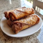 Four golden-brown, crispy peach cobbler egg rolls piled on a white plate and dusted heavily with granulated sugar.