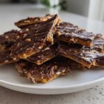 A stack of broken pieces of homemade Pecan Pie Bark topped with drizzled chocolate and nuts, served on a white plate.