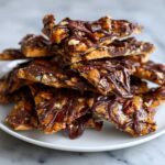 A close-up stack of broken pieces of Pecan Pie Bark drizzled with dark chocolate on a white plate.