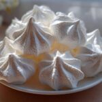 A close-up of several perfectly piped, crisp white meringue cookies piled on a small white plate.