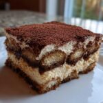 A close-up of a perfect square slice of tiramisu recipe, layered with cream and coffee-soaked ladyfingers, dusted with cocoa.