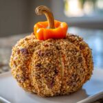 A festive pumpkin cheese ball coated in nuts and crumbs, topped with a small orange pepper stem.