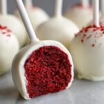 Close-up of a white chocolate-coated red velvet cake pop cut in half showing the vibrant red interior, surrounded by other red velvet cake pops.