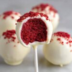 Close-up of a red velvet cake pop dipped in white chocolate, showing the rich red interior and red crumb topping.