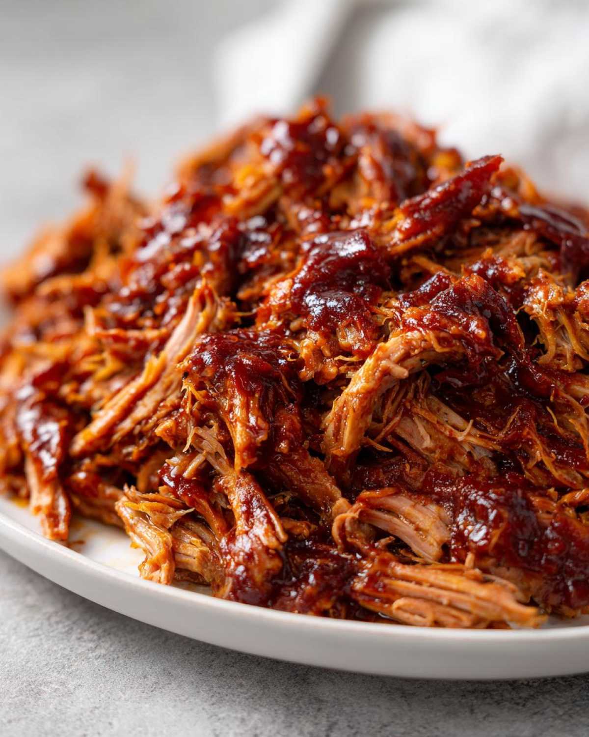 A close-up, mouthwatering shot of shredded crock pot pulled pork heavily coated in dark barbecue sauce on a white plate.