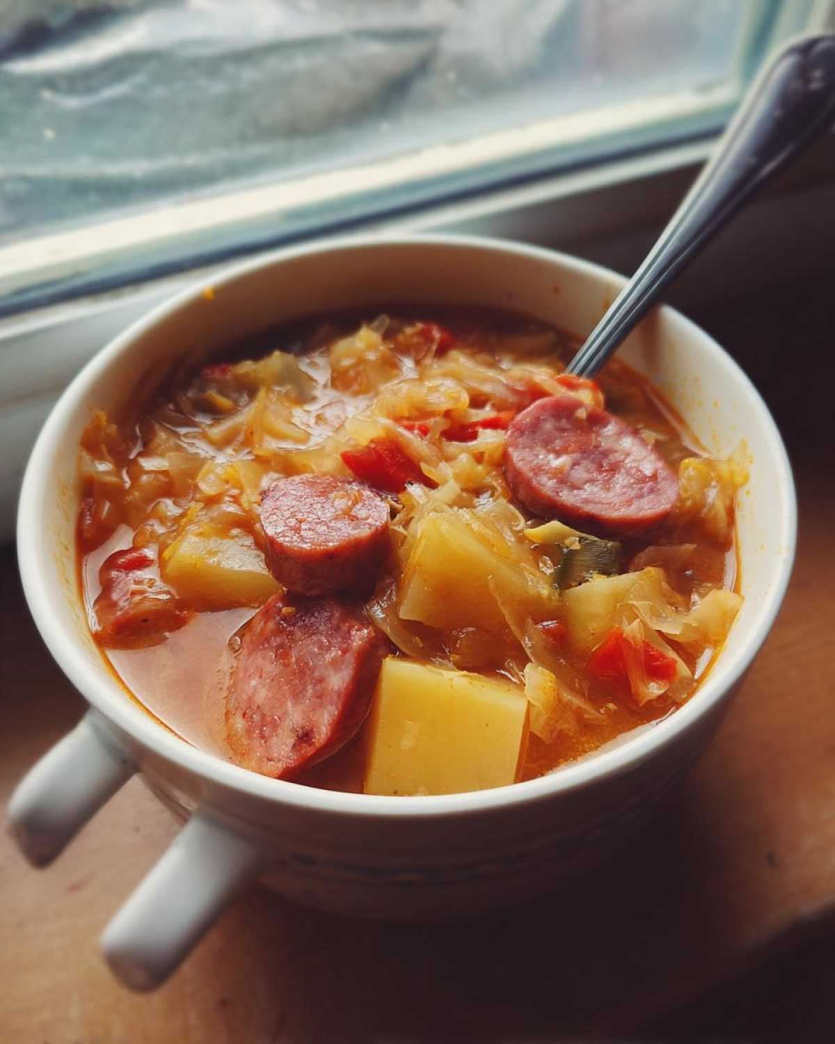 Close-up of a steaming bowl of sauerkraut soup featuring sliced sausage and chunks of potato, set near a window.