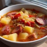 Close-up of a steaming bowl of sauerkraut soup featuring chunks of sausage, potatoes, and tomatoes.