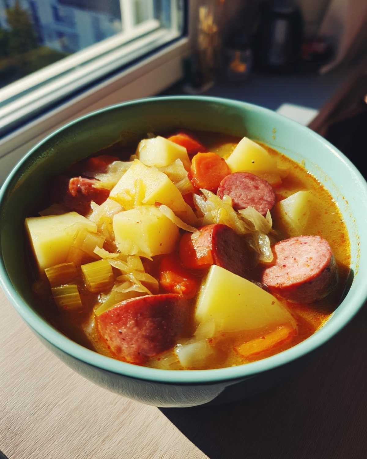 A close-up of a bowl of hearty sauerkraut soup filled with chunks of potato, sausage slices, carrots, and cabbage.