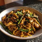 Close-up of a bowl filled with savory chicken chow mein recipe, featuring noodles, chicken pieces, and green vegetables.