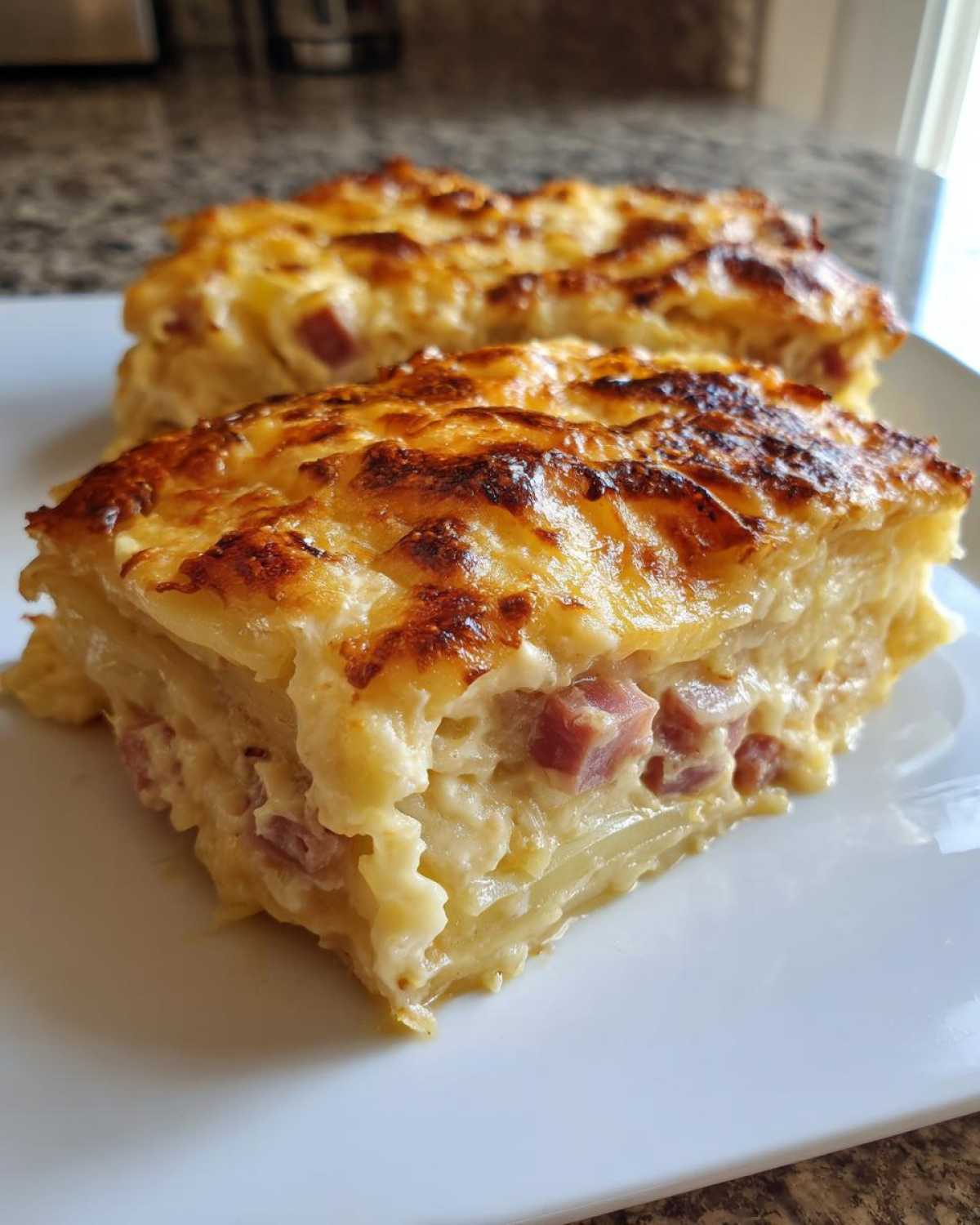 Two thick slices of creamy scalloped potatoes and ham with a bubbly, browned cheese topping served on a white plate.