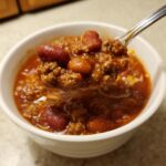 A spoonful of rich, meaty crockpot chili, featuring kidney beans and ground beef, lifted from a white bowl.